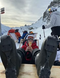 a man sitting on top of a wooden bench eating an ice cream cone with skis in the background