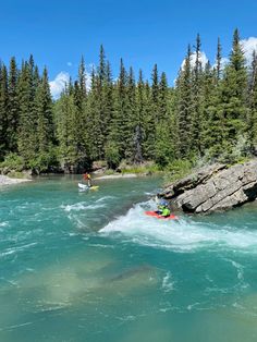 two people are kayaking in the water near some rocks and pine trees, while another person is on a raft behind them