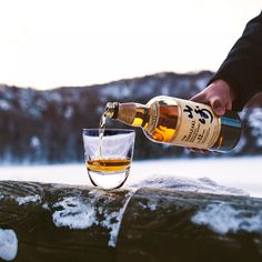 a person pouring whiskey into a glass on top of snow covered ground with mountains in the background