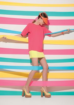 a woman in pink shirt and yellow skirt with colorful strips on the wall behind her