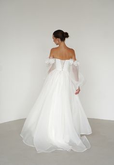 a woman in a white wedding dress with long sleeves and flowers on the shoulder is looking back