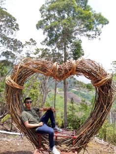 a man sitting in a heart shaped chair made out of sticks and branches with trees behind him