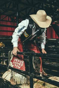 a man with a cowboy hat on leaning over a fence holding a bag and looking at the ground