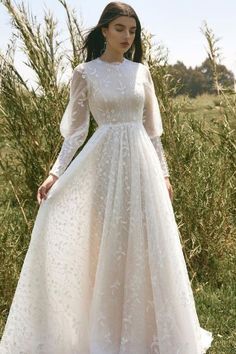 a woman wearing a white dress standing in front of some tall grass and bushes with her hands on her hips