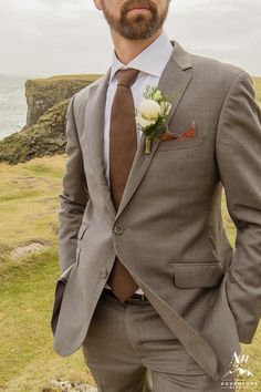 a man wearing a suit and tie standing in front of the ocean with his hands in his pockets