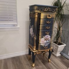 an ornate black and gold chest of drawers with flowers painted on the front, sitting next to a potted plant