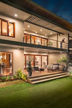 a man standing on the balcony of a modern house at night with lights on and green grass in front