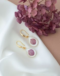 two pairs of earrings sitting on top of a white cloth next to purple flowers and greenery