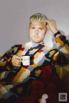 a man sitting on a bed holding a coffee cup in his hand and looking at the camera