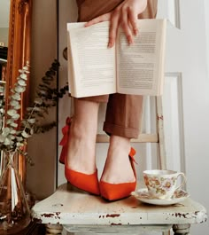 a woman sitting on a chair reading a book and holding a cup with her legs