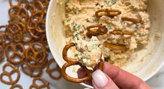 a person holding a pretzel in front of a bowl of dip and pretzels