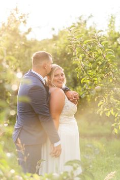 a bride and groom standing in an apple orchard with the sun shining through the trees