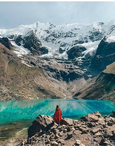 a person sitting on top of a mountain next to a lake with snow covered mountains in the background