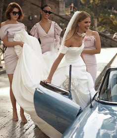 three beautiful women in dresses walking next to a blue car and one is carrying a bride's dress