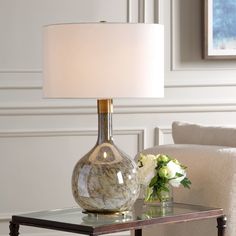 a table with a lamp and flowers on it in front of a white chair next to a couch