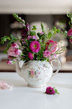 a white tea pot filled with pink flowers on top of a table next to another vase