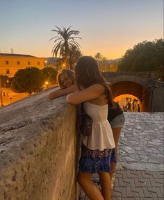 two girls leaning against a wall with their arms around each other as the sun sets