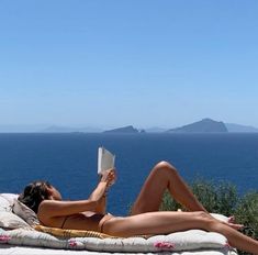 a woman laying on top of a beach next to the ocean with a book in her hand