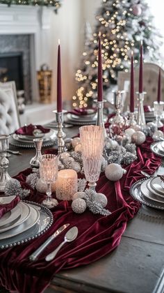 the table is set for christmas dinner with silver and red decorations on it, along with candles