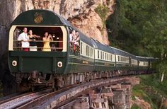 a green and yellow train traveling down tracks next to a rocky mountain side with people on it