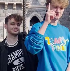 two young men standing next to each other in front of a castle with one pointing at the camera