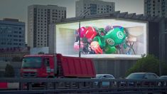 a red truck driving past a large billboard on the side of a road in front of tall buildings