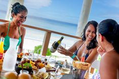 three women sitting at a table with food and drinks in front of them, one holding a bottle of wine