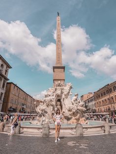 a woman standing in front of a fountain surrounded by tall buildings and people walking around