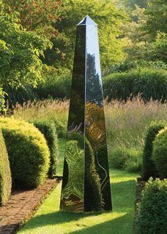 a sculpture in the middle of a garden surrounded by trees and bushes, with an open field behind it