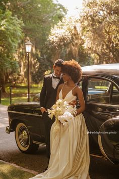 a bride and groom standing in front of an old black car on the side of the road