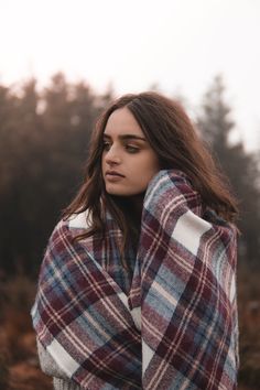 a woman wrapped in a plaid blanket looking off into the distance with trees in the background