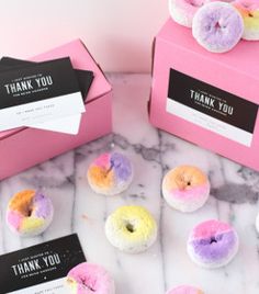 several donuts with different colored frosting are on a marble countertop next to pink boxes that say thank you