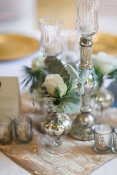 a table topped with vases filled with white flowers and greenery next to candles