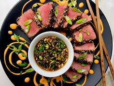 a black plate topped with slices of meat next to chopsticks and a bowl of sauce