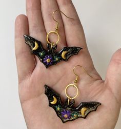 two bat shaped earrings in the palm of someone's hand, with flowers on them