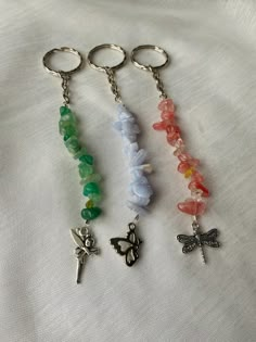 three key chains with charms attached to them on a white cloth covered surface, one has a butterfly and the other has a cross