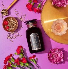 a bottle of black liquid next to some flowers on a purple surface with a gold plate