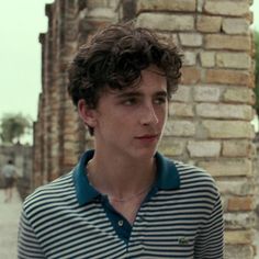 a man with curly hair wearing a blue and white striped shirt standing in front of a brick wall