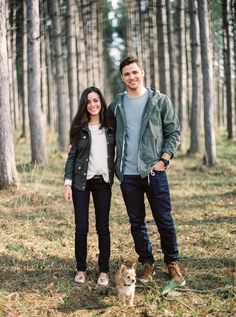 a man and woman standing next to each other in the woods with a small dog