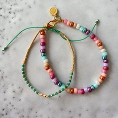 two bracelets with colorful beads are on a marble surface, one has a gold charm and the other is a green string