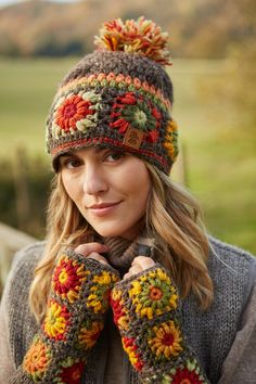 a woman wearing a knitted hat and mittens with flowers on the top, standing in front of a green field