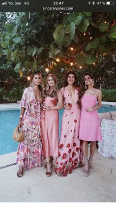 four women standing next to each other in front of a pool with lights strung overhead