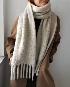 a woman wearing a white scarf with fringes on the ends is standing in front of a wall