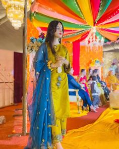 a woman dressed in yellow and blue standing under a colorful tent with people sitting around it