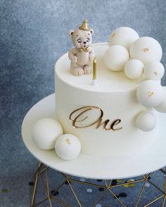 a cake decorated with white balls and a small teddy bear on top that says one