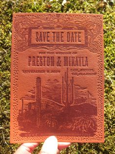 a person holding up a leather save the date card in front of some green grass