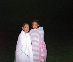 two women wrapped up in towels standing next to each other on the beach at night