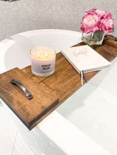 a wooden tray with a candle and notebook on it next to a pink flower in a glass vase