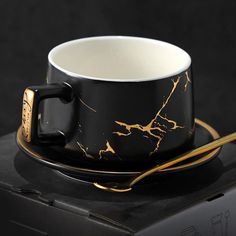 a black and gold coffee cup sitting on top of a box next to a golden spoon