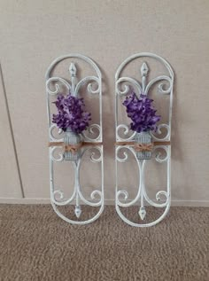 two white iron wall shelves with purple flowers in them on carpeted floor next to beige wall